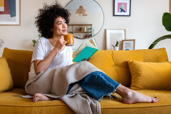 woman sits on sofa with a book, blanket, mug